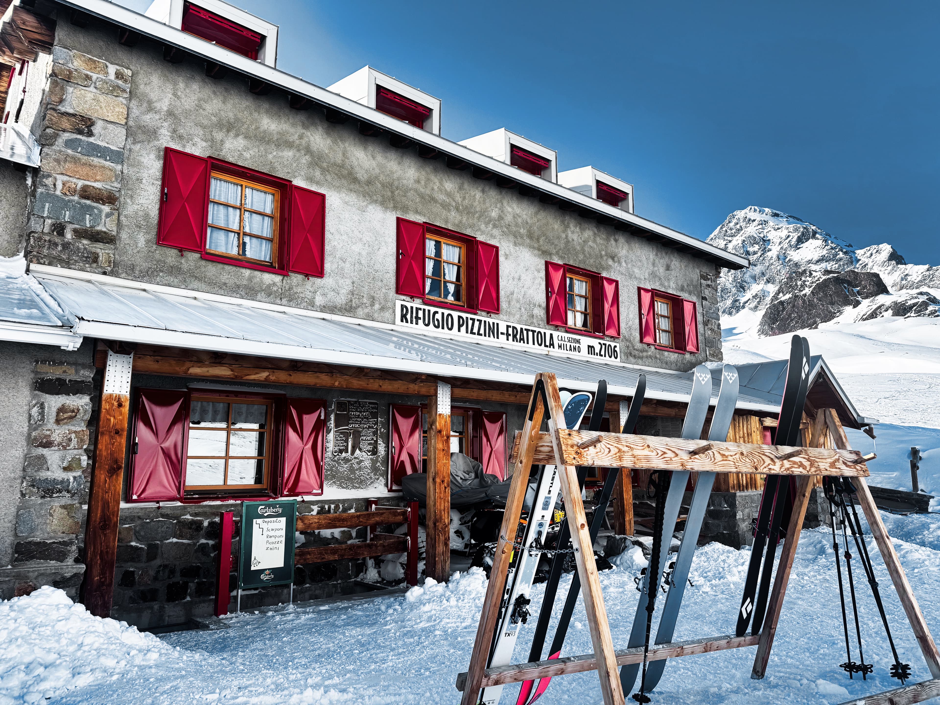 Rifugio Pizzini-Frattola at 2,706m, skis racked outside, a peak looming behind