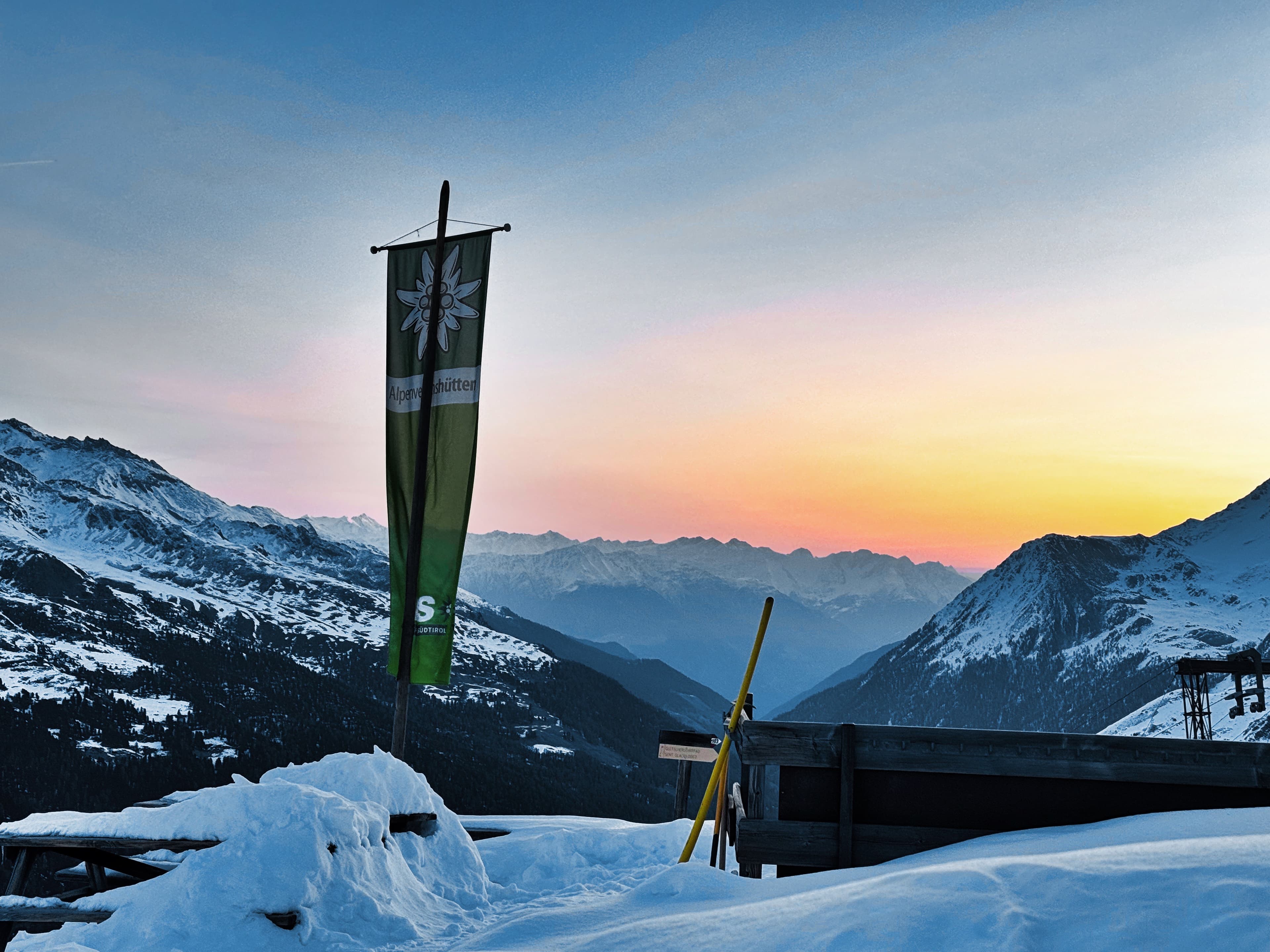 Sunset over the valley from the Branco hut deck, Alpenvereins flag, a single ski propped against the railing